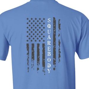 May include: Sky blue t-shirt featuring a distressed American flag design on the back. The flag is composed of stars and stripes in shades of gray, with the word "SQUAREBODY" printed vertically alongside the flag.