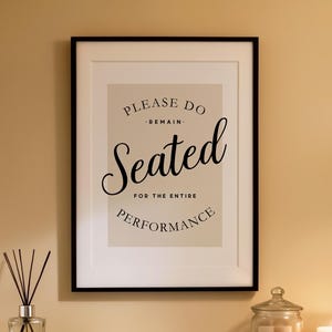 Può includere: Una stampa incorniciata con la scritta "PLEASE DO REMAIN SEATED FOR THE ENTIRE PERFORMANCE" è appesa sopra una mensola con una candela, un barattolo e degli asciugamani. L'arredamento del bagno comprende un water e un diffusore a bastoncini.