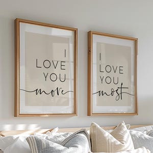 Op de afbeelding: Twee ingelijste prints met de zinnen "I Love You More" en "I Love You Most" in zwart schrift. De prints worden weergegeven boven een bed met wit beddengoed en decoratieve kussens. De frames zijn lichtbruin.