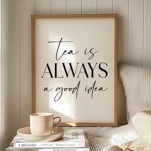 May include: A framed print with the text "Tea is ALWAYS a good idea" in black script. A beige teacup and saucer sit atop a stack of books. The scene is set in a bright, cozy room with a window.