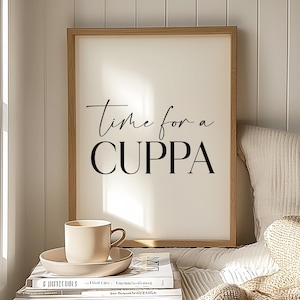 Puede incluir: Una impresión enmarcada con el texto "Time for a CUPPA" en escritura negra. Una taza y un platillo beige están sobre una pila de libros. La escena está bañada en luz suave, sugiriendo un ambiente acogedor. La impresión está en un marco de madera.