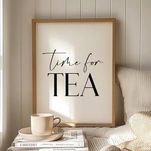May include: A framed print with the words "Time for TEA" in black script and block letters. A beige teacup and saucer sit atop a stack of books. The scene is set in a bright, cozy room with a window and a bed.