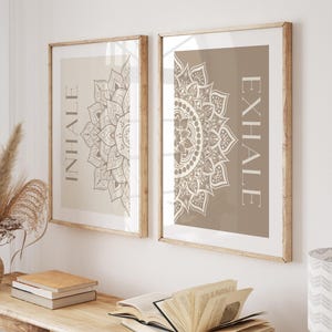 May include: Two framed art prints with the words "INHALE" and "EXHALE" and a mandala design. The prints are in a room setting with a wooden table, books, and a vase of dried flowers. The prints are in wooden frames.