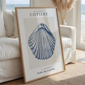 May include: A framed vertical wall art print featuring a detailed blue line drawing of a seashell centred on a light beige background. The text EXPOSITION DE CÔTIÈRE appears at the top, with EN VACANCES Another day in paradise at the bottom.