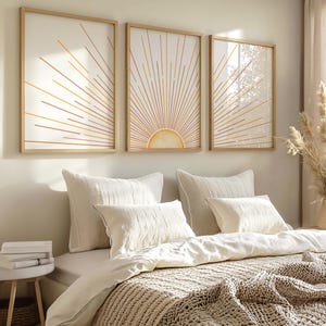 Könnte beinhalten: Drei gerahmte Wandkunstwerke mit einem Sonnenstrahl-Design in Gold- und Orangetönen. Die Kunstwerke hängen über einem Bett mit weißen Kissen und einer beigefarbenen Strickdecke. Der Text auf der linken Seite lautet "PROFESSIONAL GRADE - PREMIUM SATIN PHOTOPAPER".