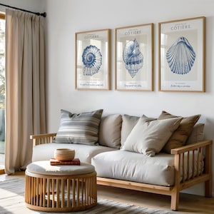 K&ouml;nnte beinhalten: Drei gerahmte Drucke mit blauen und wei&szlig;en Illustrationen von Muscheln. Die Drucke h&auml;ngen an einer wei&szlig;en Wand &uuml;ber einem hellbeigen Sofa mit Kissen. Das Sofa ist aus Holz und hat ein hellbeiges Stoffkissen.