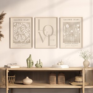 May include: Three beige framed prints with white botanical illustrations. The left print features leaves and the text "Galerie d'Art Exposition Botanique Feuilles". The centre print features the word "LOVE" in large letters. The right print features flowers and the text "Galerie d'Art Exposition Botanique Jardin de Fleurs".