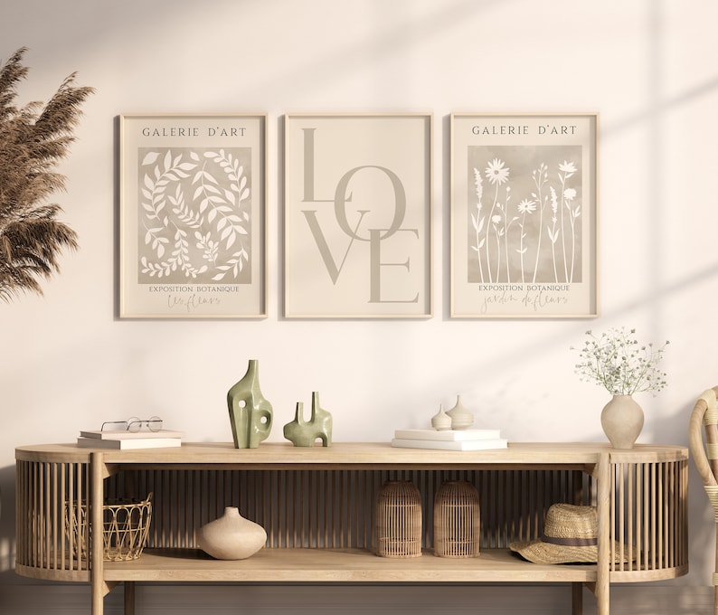 May include: Three beige framed prints with white botanical illustrations. The left print features leaves and the text "Galerie d'Art Exposition Botanique Feuilles". The centre print features the word "LOVE" in large letters. The right print features flowers and the text "Galerie d'Art Exposition Botanique Jardin de Fleurs".