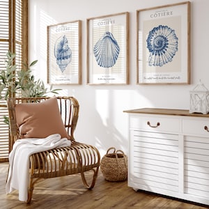 K&ouml;nnte beinhalten: Drei gerahmte Drucke mit blauen Illustrationen von Muscheln auf cremefarbenem Hintergrund. Die Drucke sind mit "Cottiere" betitelt und zeigen den Text "Every sunset may be a dream", "Another day in paradise" und "The perfect hearts build the hardest shells".