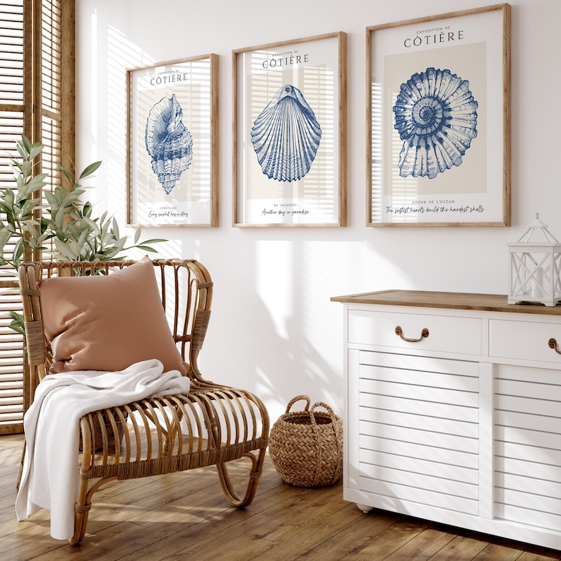 K&ouml;nnte beinhalten: Drei gerahmte Drucke mit blauen Illustrationen von Muscheln auf cremefarbenem Hintergrund. Die Drucke sind mit "Cottiere" betitelt und zeigen den Text "Every sunset may be a dream", "Another day in paradise" und "The perfect hearts build the hardest shells".