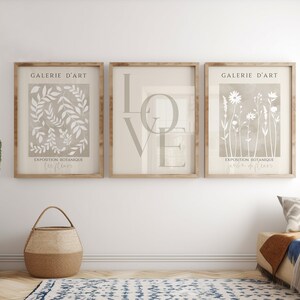 May include: Three framed prints with a beige background and white designs. The left print features a botanical illustration of leaves and the text "EXPOSITION BOTANIQUE JARDIN DE FLEURS". The middle print features the word "LOVE" in large, stylized letters. The right print features a botanical illustration of flowers and the text "EXPOSITION BOTANIQUE JARDIN DE FLEURS". All three prints have the text "GALERIE D'ART" at the top.