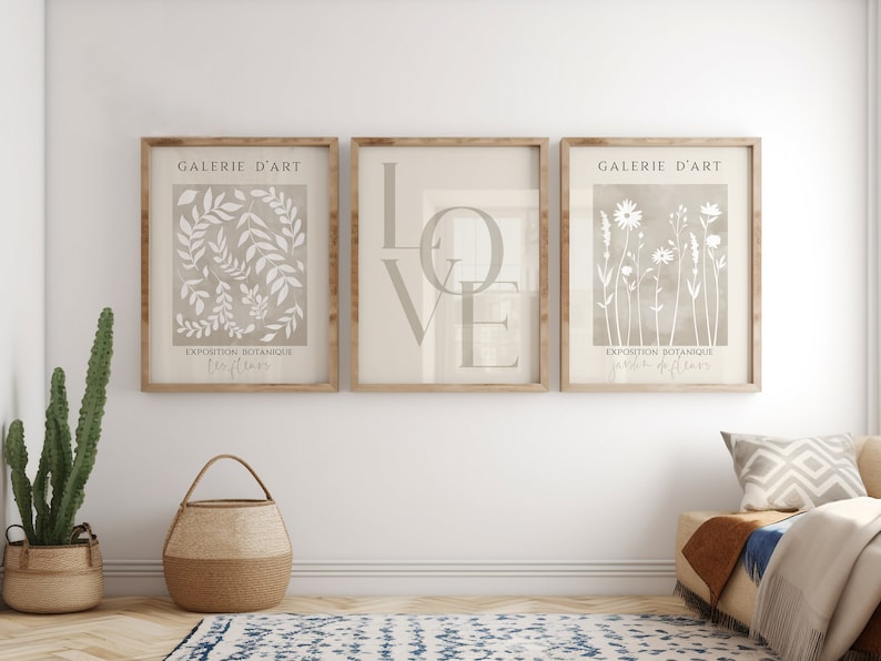 May include: Three framed prints with a beige background and white designs. The left print features a botanical illustration of leaves and the text "EXPOSITION BOTANIQUE JARDIN DE FLEURS". The middle print features the word "LOVE" in large, stylized letters. The right print features a botanical illustration of flowers and the text "EXPOSITION BOTANIQUE JARDIN DE FLEURS". All three prints have the text "GALERIE D'ART" at the top.