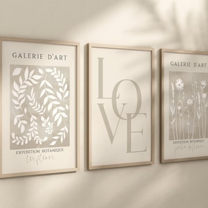 May include: Three framed prints with a beige background and white illustrations. The left print features a botanical illustration of leaves and the text "Galerie d'Art Exposition Botanique Les Fleurs". The middle print features the word "LOVE" in large, stylized letters. The right print features a botanical illustration of flowers and the text "Galerie d'Art Exposition Botanique Jardin de Fleurs".