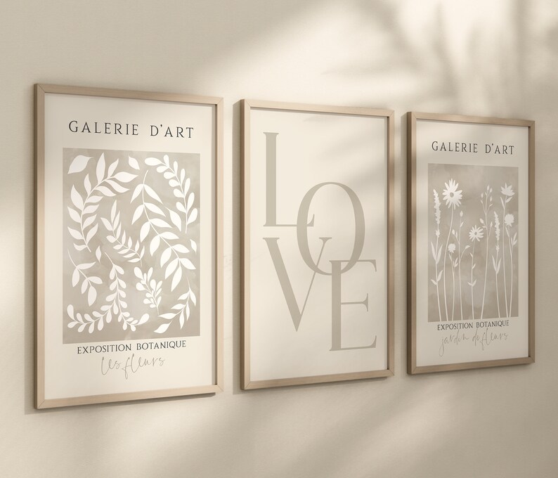 May include: Three framed prints with a beige background and white illustrations. The left print features a botanical illustration of leaves and the text "Galerie d'Art Exposition Botanique Les Fleurs". The middle print features the word "LOVE" in large, stylized letters. The right print features a botanical illustration of flowers and the text "Galerie d'Art Exposition Botanique Jardin de Fleurs".