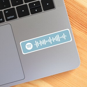 Peut inclure: Un autocollant rectangulaire sur fond bleu clair avec le logo Spotify blanc sur la gauche et un motif de code d'onde sonore blanc s'&eacute;tendant vers la droite. L'autocollant est appos&eacute; sur la surface m&eacute;tallique d'un ordinateur portable.