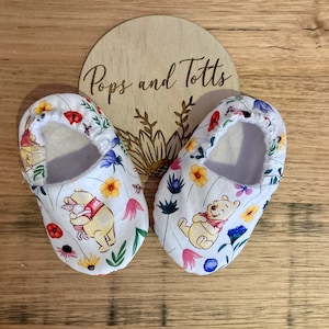 May include: Baby shoes with a white floral pattern and Winnie the Pooh illustrations. The shoes are on a wooden surface, with a wooden sign that reads "Pops and Totts". The shoes have a soft, elasticated opening.