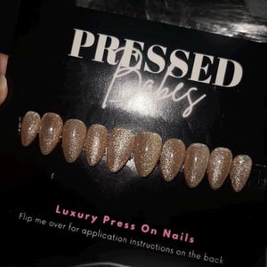 May include: A set of gold glitter press-on nails. The packaging reads "PRESSED Babes" in white script. The text "Luxury Press On Nails" is also visible. The image also includes the text "Flip me over for application instructions on the back."