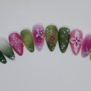 May include: Ten long, oval-shaped artificial nails with a gradient design. The nails are painted with shades of green, white, and pink. Some nails have a floral design with white flowers and green leaves.