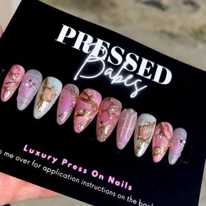 May include: A set of ten pink and white press-on nails with gold accents and pearl embellishments. The nails are displayed on a black card with the text "Pressed Babes" and "Luxury Press On Nails".