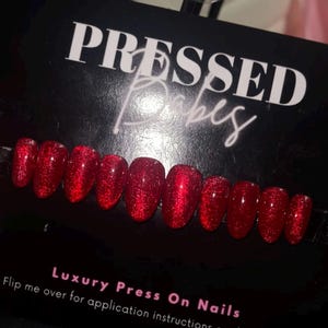 May include: A set of red glitter press-on nails. The nails are oval-shaped and have a shiny, textured appearance. The product is displayed on a black card with the text "PRESSED Babes" and "Luxury Press On Nails."