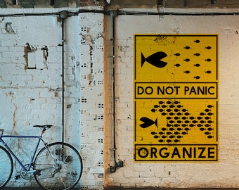 May include: A yellow rectangular poster on a white brick wall featuring black graphics of small fish forming a larger fish shape. The text reads DO NOT PANIC in the centre and ORGANIZE at the bottom. A blue bicycle is positioned to the left.