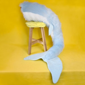 May include: A plush, light blue whale tail prop with a white belly and a large, fan-shaped tail. The tail is draped on a yellow stool with a yellow cushion, set against a bright yellow backdrop. The prop is made of soft, furry material.