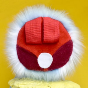 May include: A colorful, whimsical hat with a red top and a white fur trim. The hat features a red ribbon detail and a white circular accent. The hat is set against a yellow background.