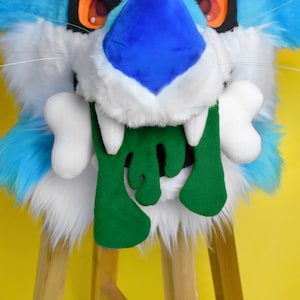May include: A furry creature mask in shades of blue, white, and turquoise, featuring large orange eyes and white teeth. The mask has a green, drippy, felt-like design from the mouth and two white bone-shaped accents.
