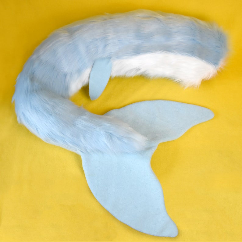 May include: A light blue and white faux fur tail with a matching light blue fin. The tail is fluffy and curved, with a smooth, flat fin at the end. The tail is designed to be worn as a costume accessory.