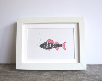 May include: Framed art print with a stylised fish illustration. The fish is black with red fins and tail, and has three small blue bubbles. The artwork is set in a white rectangular frame, suitable for a home.