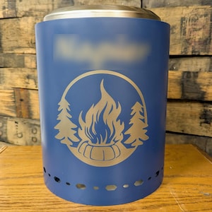 May include: A blue metal fire pit with a gold-colored campfire graphic. The graphic includes a campfire, trees, and a circular border. The fire pit has a silver top and small holes around the base.