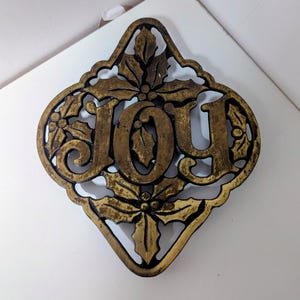 May include: A decorative metal sign in a diamond shape, featuring the word "JOY" in large, ornate letters. The sign is gold-colored with black accents and holly leaf designs, perfect for holiday decor.