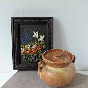 May include: A round, glazed ceramic pot with two side handles and a matching lid, featuring a gradient of tan and brown tones. Beside it stands a rectangular black frame containing a textured oil painting of colorful flowers.