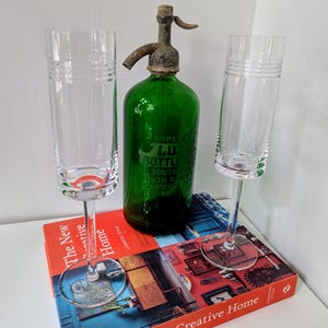 May include: A green glass seltzer bottle with a metal top is flanked by two clear champagne flutes. The flutes and bottle sit on a red book titled "The New Creative Home". The bottle has text that reads "ROPE LU BOTTL SOUTH SOUTH R CONTENU".