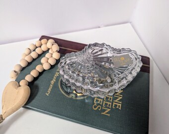Può includere: Un portagioie di cristallo trasparente a forma di cuore con un design strutturato è appoggiato su un libro verde scuro. Una ghirlanda di perline di legno con un ciondolo a forma di cuore è accanto.