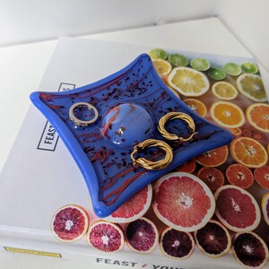 Op de afbeelding: Een blauwe glazen schaal met een rood en wit swirl-ontwerp. De schaal staat op een boek met een kleurrijk fruitpatroon. Er zitten twee gouden oorringen en een zilveren ring in de schaal.