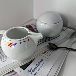 Peut inclure: Une crémier en céramique blanche avec un motif coloré et un sucrier en céramique blanche avec un couvercle. La crémier a une poignée et un bec verseur. Le sucrier a un couvercle avec un bouton. Les deux pièces sont sur un journal avec une cuillère.