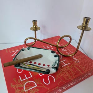 Könnte beinhalten: Ein Keramik-Aschenbecher mit einer Zigarre, einem Messing-Kerzenhalter und einem roten Buch. Der Aschenbecher ist weiß mit einem farbenfrohen Rand und dem Wort "Hatia". Der Kerzenhalter hat zwei Kerzenschalen.