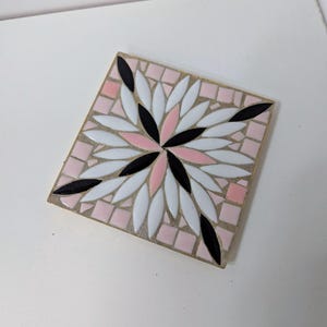 Puede incluir: Azulejo de mosaico cuadrado con diseño floral. El azulejo presenta un patrón de piezas en forma de pétalo blancas, negras y rosas dispuestas alrededor de un punto central. El fondo es un mosaico rosa claro.