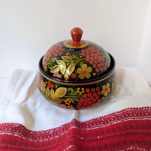 May include: A black, round, lidded bowl with a vibrant floral design. The bowl features red berries, yellow flowers, and gold leaves against a black background. The lid has a small, red knob. The bowl sits on a white cloth with a red and white patterned border.