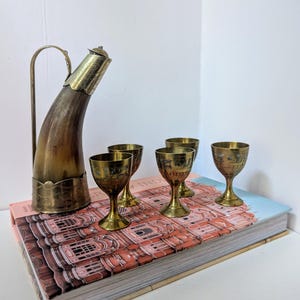 Puede incluir: Un juego de cinco vasos para beber de latón y una jarra de licor de latón y cuerno. Los vasos tienen diseños intrincados y la jarra tiene un pico de cuerno.
