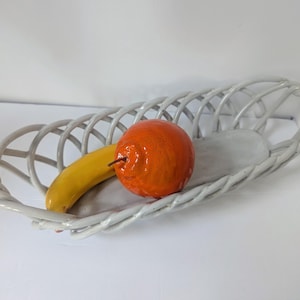 May include: A white, oval-shaped woven basket holds a yellow banana and an orange fruit. The basket has a braided rim and a solid, light gray base. The fruit is placed on the gray base.