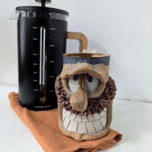 May include: A black French press with a glass carafe and a light wood handle, positioned next to a handmade ceramic mug featuring a sculpted face with a large nose, textured brown beard, and a wide, toothy grin under a dark brown brim.