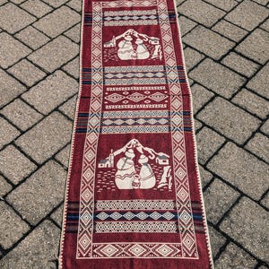 May include: A red and white woven rug with a geometric pattern and a design of two figures in traditional clothing. The rug is laid out on a gray brick surface.