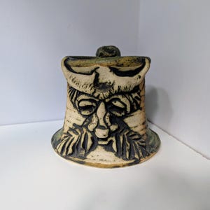 Puede incluir: Una escultura de cerámica en forma de campana con un diseño de rostro tallado y follaje. La escultura es principalmente beige con detalles en marrón oscuro y verde. La cara tiene una nariz y ojos prominentes, con un pequeño pomo en la parte superior.