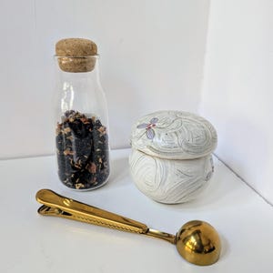 May include: A set of tea accessories including a glass jar with a cork lid filled with loose leaf tea, a white ceramic tea caddy with a swirling design and a gold metal tea scoop with a clip.