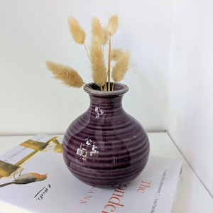 Puede incluir: Un jarrón de cerámica de color morado oscuro brillante con cuerpo redondeado y textura acanalada horizontal. El jarrón tiene un cuello estrecho y un borde ensanchado, conteniendo varios tallos de hierba seca, beige y esponjosos.