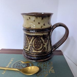 May include: A ceramic mug with a brown handle and a decorative pattern. The mug has a beige top with brown speckles and a dark brown band. The body of the mug features a repeating brown pattern. A gold spoon rests on a book.