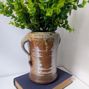 May include: A handcrafted ceramic vase with a brown and tan glaze, featuring two handles. The vase holds vibrant green artificial foliage. It sits atop a closed, navy blue book. The vase is approximately 6 inches tall.