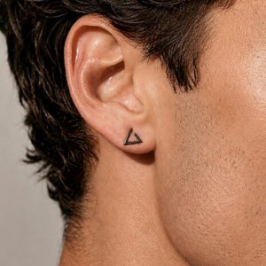 May include: A small, dark-toned metal stud earring shaped like an open triangle. The earring is positioned in the earlobe, featuring a clean, geometric outline with sharp corners and a smooth, matte finish that contrasts against the skin.